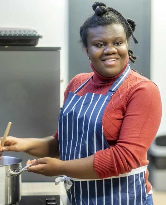 Young woman stirring a pot on the stove and smiling, wearing a striped apron in a kitchen setting. Young woman stirring a pot on the stove and smiling, wearing a striped apron in a kitchen setting.